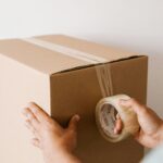 Crop anonymous male using tape to seal packed cardboard box against white wall while moving into new place