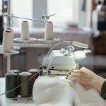 A person sewing fabric on a professional machine in a well-lit workshop with spools of thread.